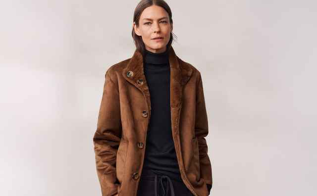 Eine Frau trägt eine braune Jacke und schwarzen Rollkragenpullover, mit den Händen in den Taschen vor einem neutralen Hintergrund.