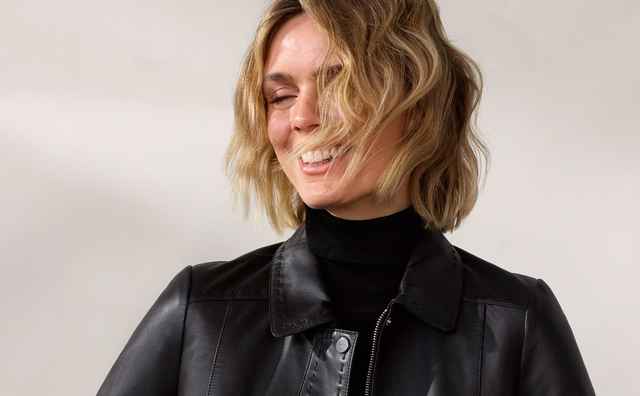 Frau lächelt und trägt eine schwarze Lederjacke über einem schwarzen Oberteil und grauen Jeans. Im Hintergrund ein Stuhl.
