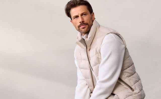Mann mit braunen Haaren trägt eine beige Steppweste und einen weißen Pullover, posierend vor neutralem Hintergrund.