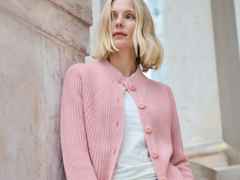 Frau mit blonden Haaren trägt eine rosa Strickjacke und ein weißes Oberteil. Sie lehnt lächelnd an einer hellen Steinwand.
