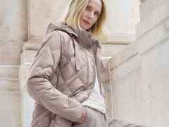 Frau in einer beigen Steppjacke posiert vor einer beigen Steinwand. Sie hat hellblondes Haar und trägt ein weißes Shirt darunter.